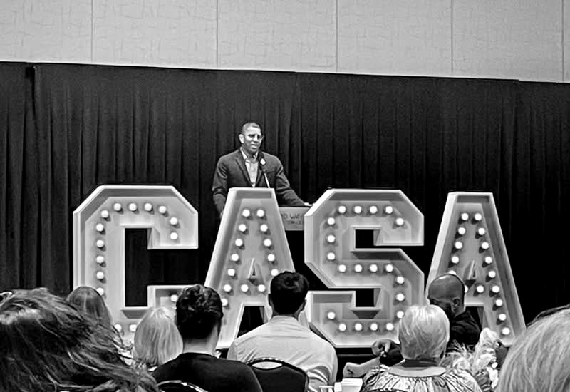 Takkeem Morgan delivering the keynote address at the 28th Annual Indiana GAL/CASA Conference in Fort Wayne, standing behind large illuminated CASA marquee letters