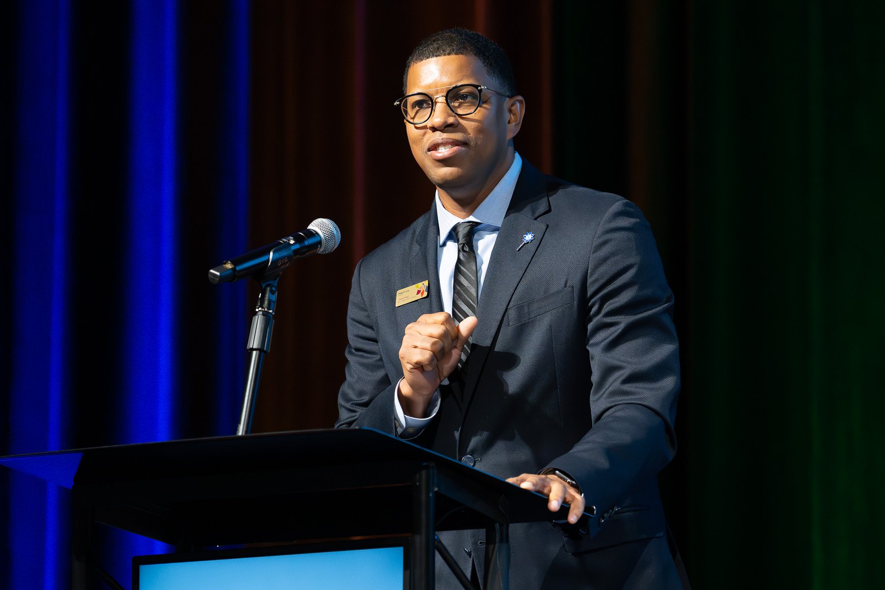 Portrait of Takkeem Morgan gesturing at the podium during formal remarks, wearing a navy suit