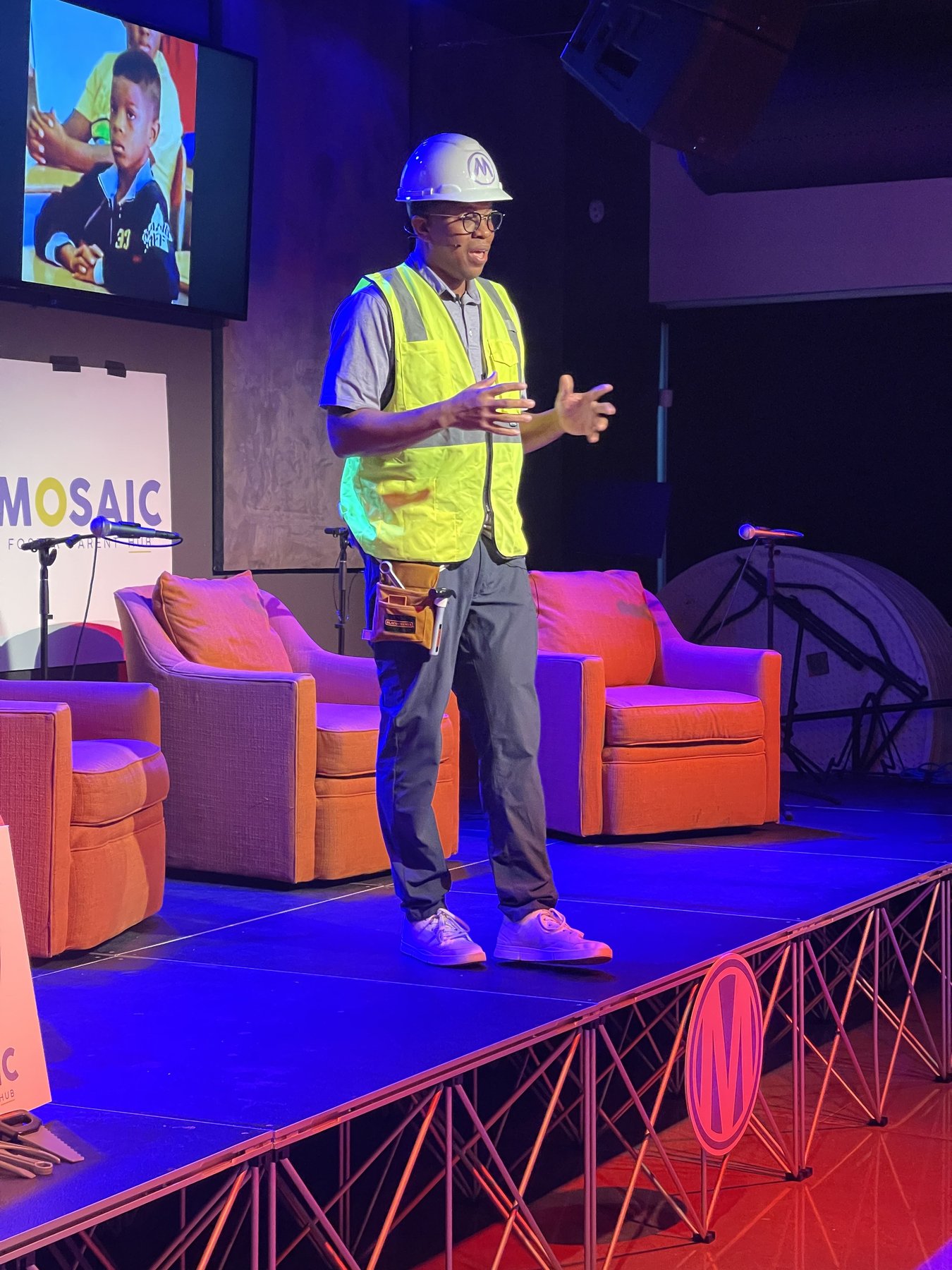 Takkeem Morgan on stage in a hard hat and reflective vest during a Mosaic conference keynote, orange lounge seating behind him