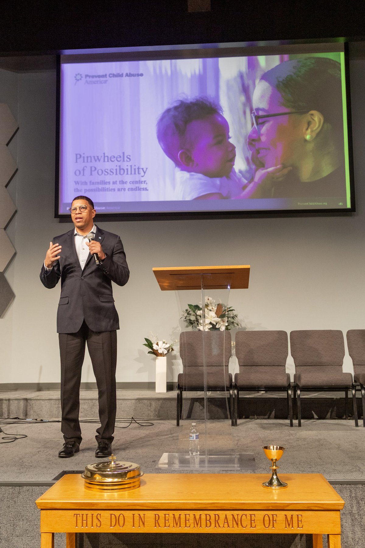 Takkeem Morgan delivering the Pinwheels of Possibility keynote, gesturing on stage in front of a large image of a mother and baby