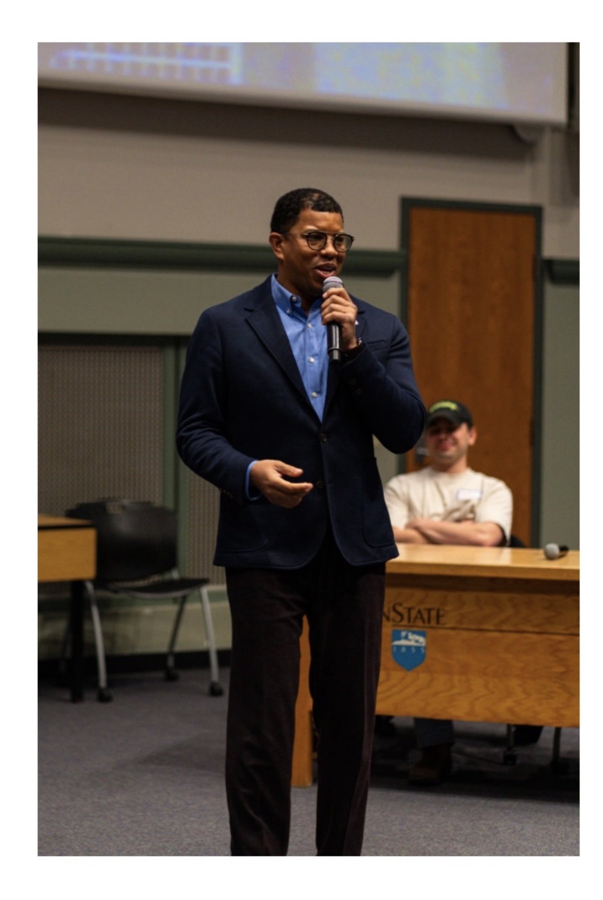 Takkeem Morgan mid-lecture at Penn State, holding a handheld microphone in a navy blazer and blue oxford shirt